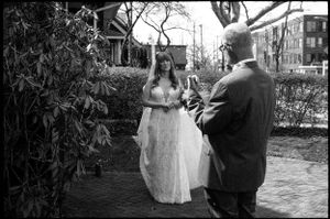 A bride in a lace gown stands outdoors with a man in a black and white photo.