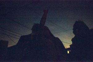 Two silhouetted figures stand outside at night under utility wires with one arm raised.