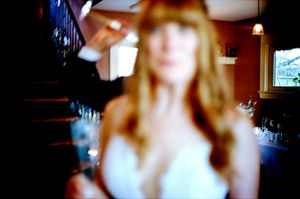 Blurred close-up of a red-haired bride in white dress holding a drink near a staircase.
