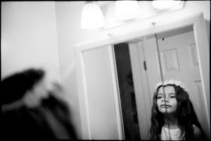A young girl with long hair and a flower crown smiles in a bathroom mirror.