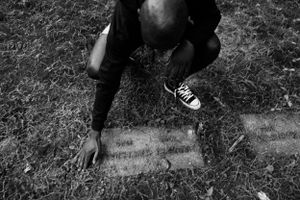 Black and white photo of person kneeling to examine old gravestone in overgrown cemetery.