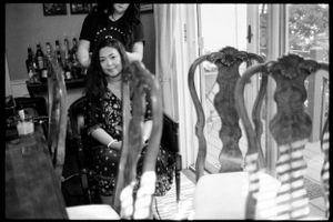 Black and white photo of a smiling Asian woman standing near ornate dining chairs in a warmly lit room.