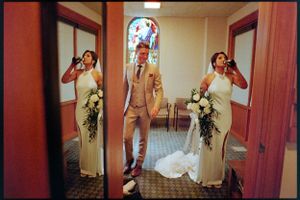 Groom stands between two bridesmaids in white dresses holding bouquets inside a church with stained glass.
