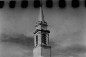 Black and white film photo of classic white church steeple against dramatic cloudy sky.