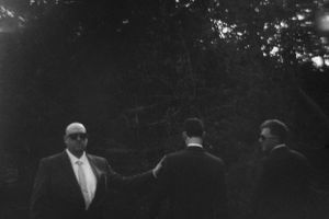 Three men in suits standing outdoors among trees in a black and white wedding photo.