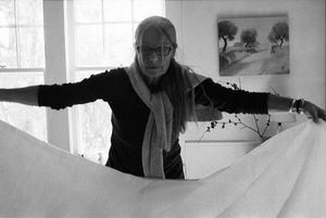Elderly woman with glasses spreading white fabric near a bright window in a black and white photo.