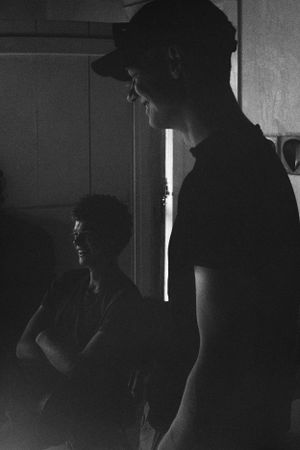 Two men in dark clothing standing in a dimly lit room with dramatic side lighting.