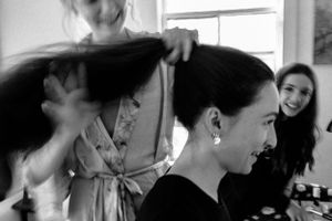 Black and white photo of woman getting hair styled while smiling friend watches in bright room.
