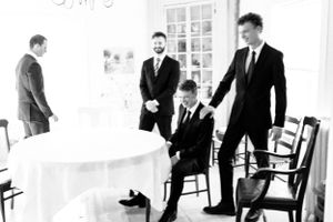 Four groomsmen in black suits gathered around a table in an elegant white room.