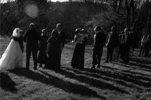 Black and white photo of wedding guests gathered outdoors on a grassy field casting long shadows.