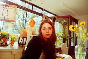 Young woman with long brown hair sits in a sunlit cafe surrounded by sunflowers and warm decor.