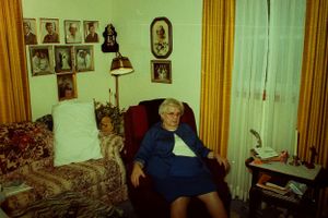 Elderly woman sitting in an armchair surrounded by family portrait photos on the wall.