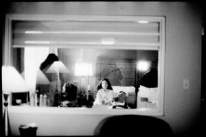 Black and white mirror reflection photo of a woman sitting in a hotel room with lamps glowing.