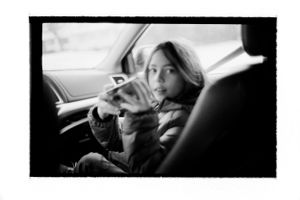 Black and white photo of a young woman applying makeup in a car.