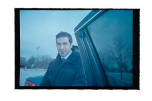 Man in dark coat standing by car door on overcast day with bare trees in background.