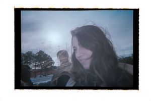 Candid film photo of a smiling woman outdoors with a blurred figure behind her.