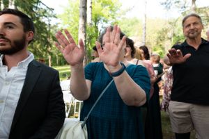 A woman raises both hands at an outdoor wedding ceremony surrounded by guests.