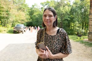Woman holding a smartphone outdoors at a sunny wedding venue with guests in the background.