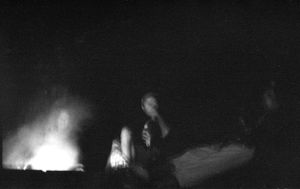 Dark black and white photo of people sitting around a glowing campfire at night.