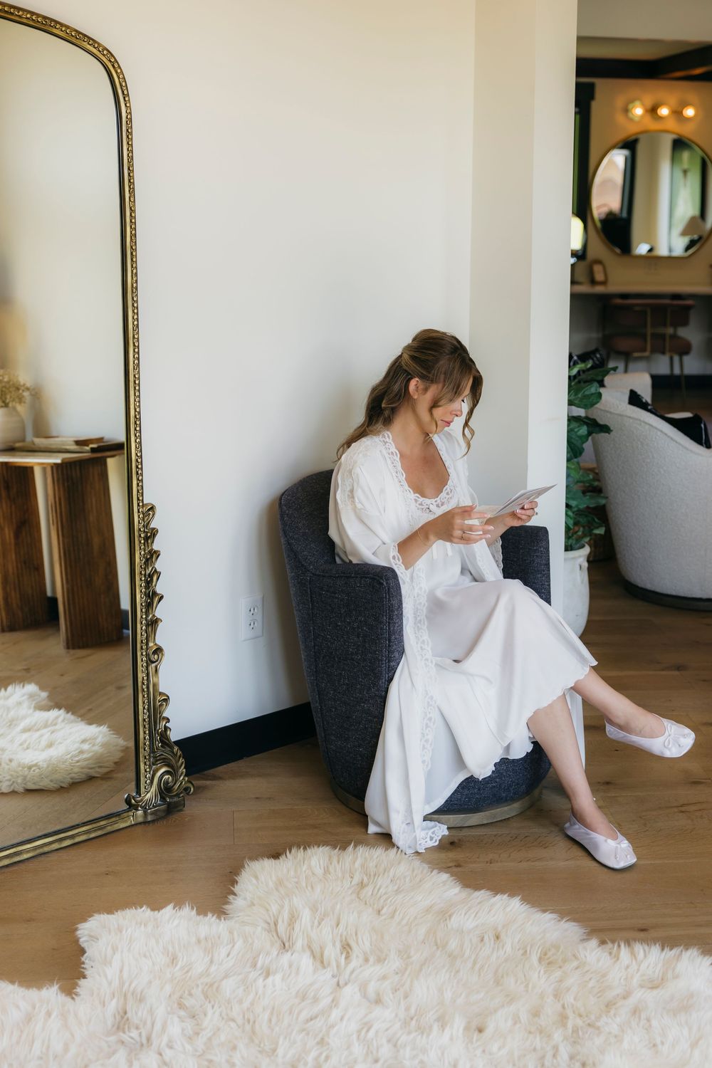 Bride in white dress and slippers reads a card while seated in a modern chair beside a gold floor mirror captured by candid photographer