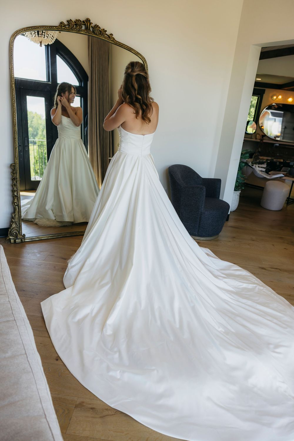 Bride in strapless ball gown with long train stands before a gold ornate mirror in a bright room captured by candid  Wisconsin photographer