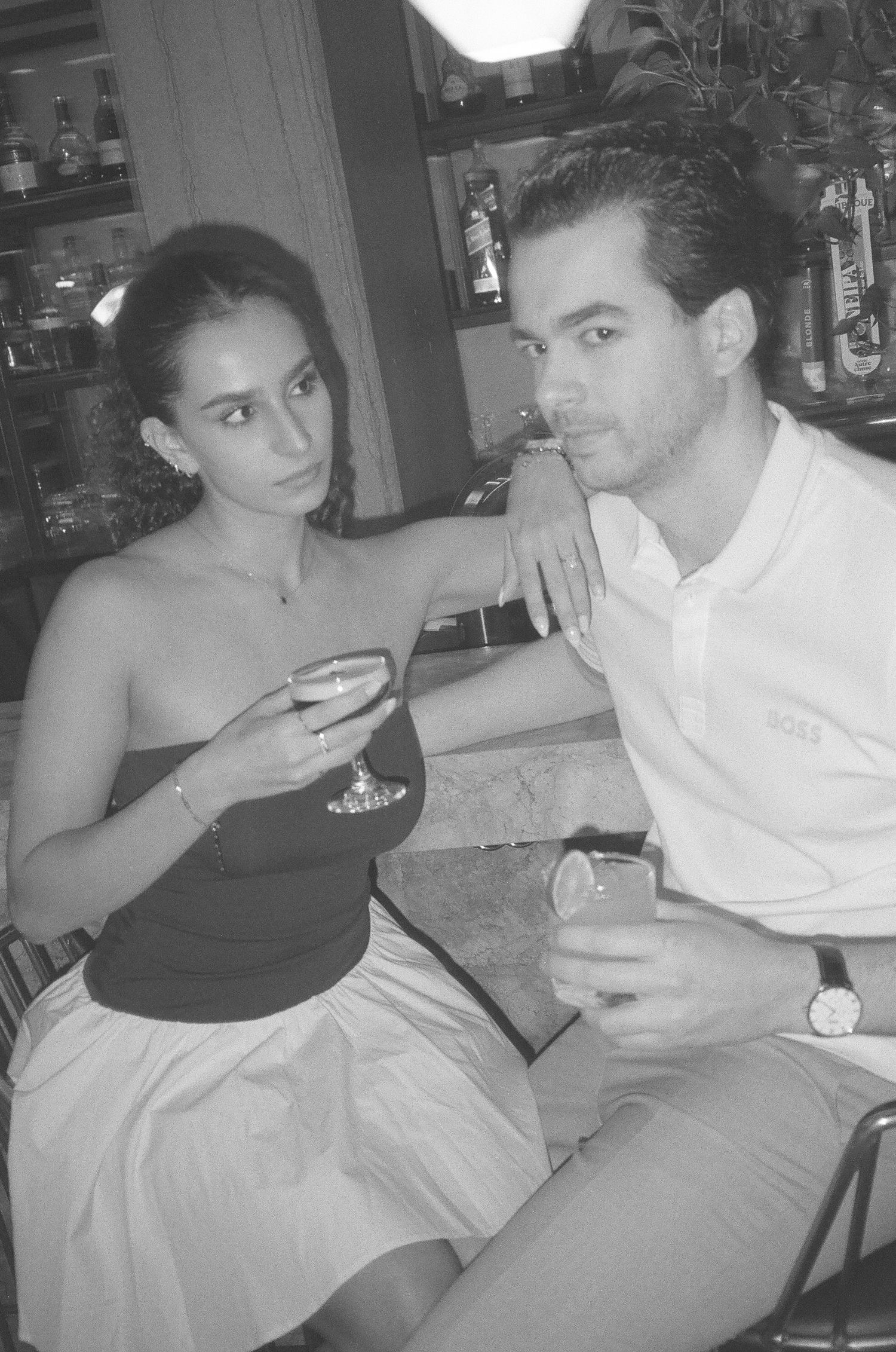 Black and white photo of a couple sitting at a bar, holding cocktail glasses and posing for the camera. Aliza Dworkind engagement photoshoot