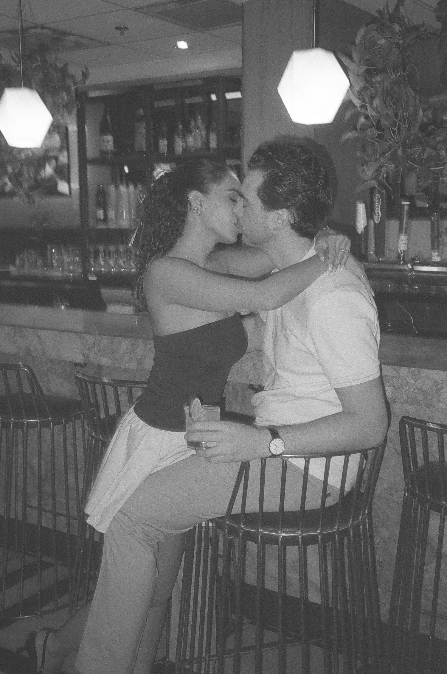 Couple sharing a romantic kiss at a bar, black and white photo, woman in black top sitting on mans lap. 35mm film photoshoot