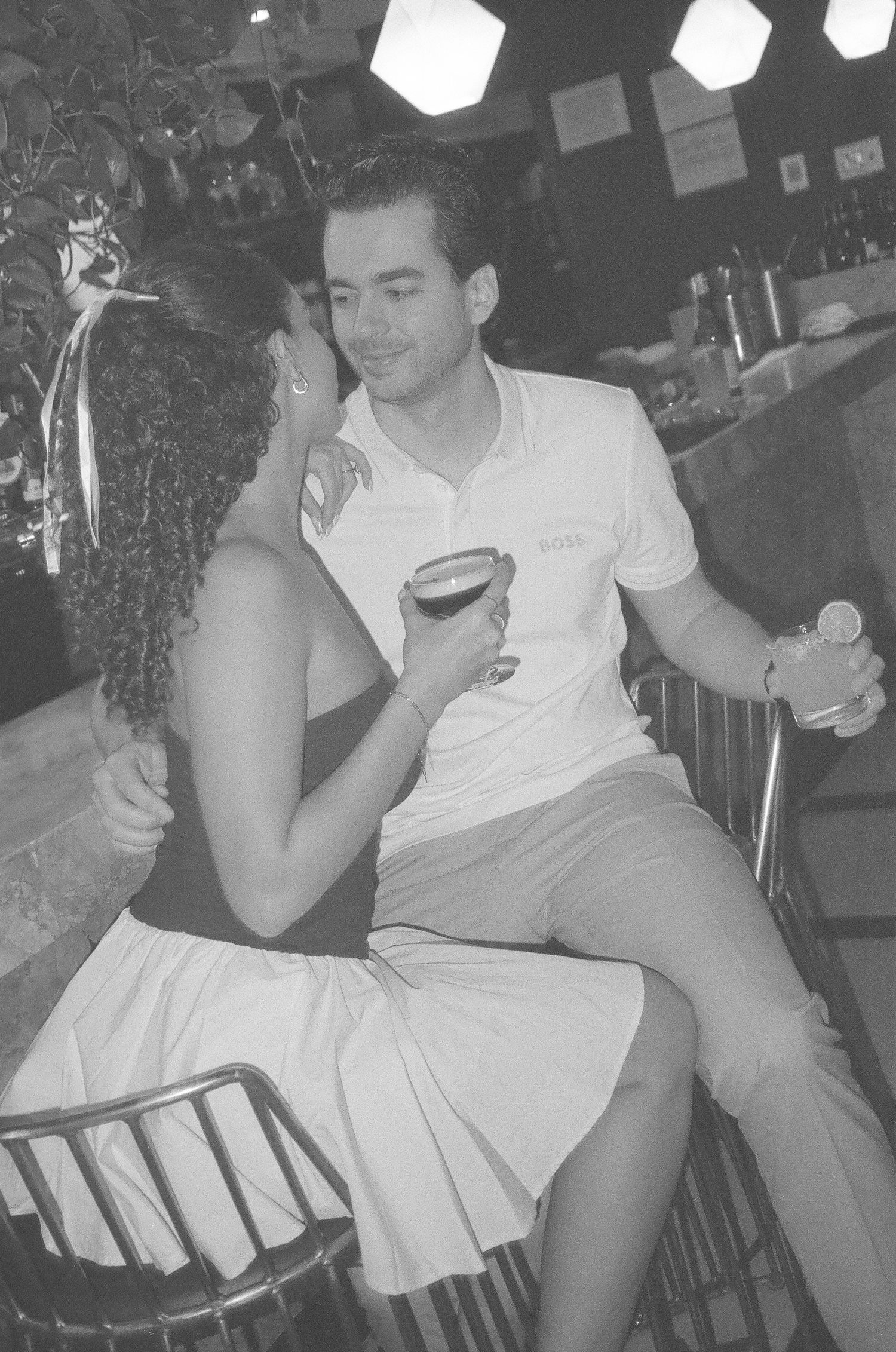 Black and white photo of a couple sharing drinks at a bar, woman in white skirt leans close to man in polo shirt. Engagement photoshoot film