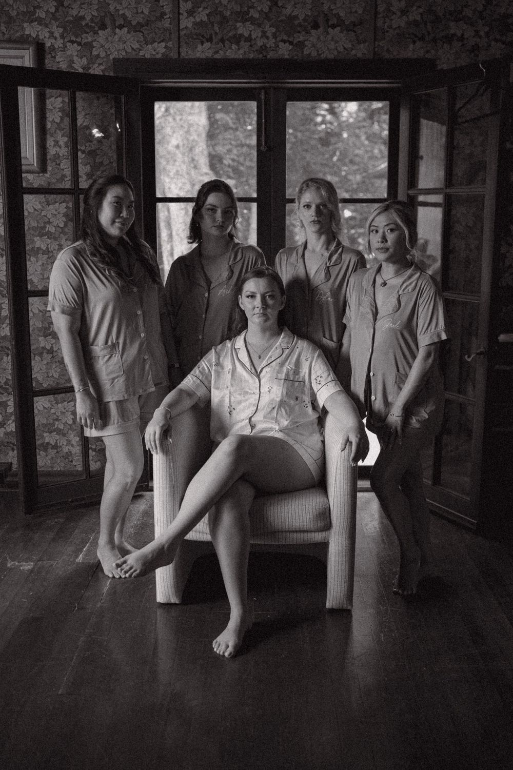 Black and white photo of bride seated in chair surrounded by four bridesmaids in matching pajamas in elegant room.