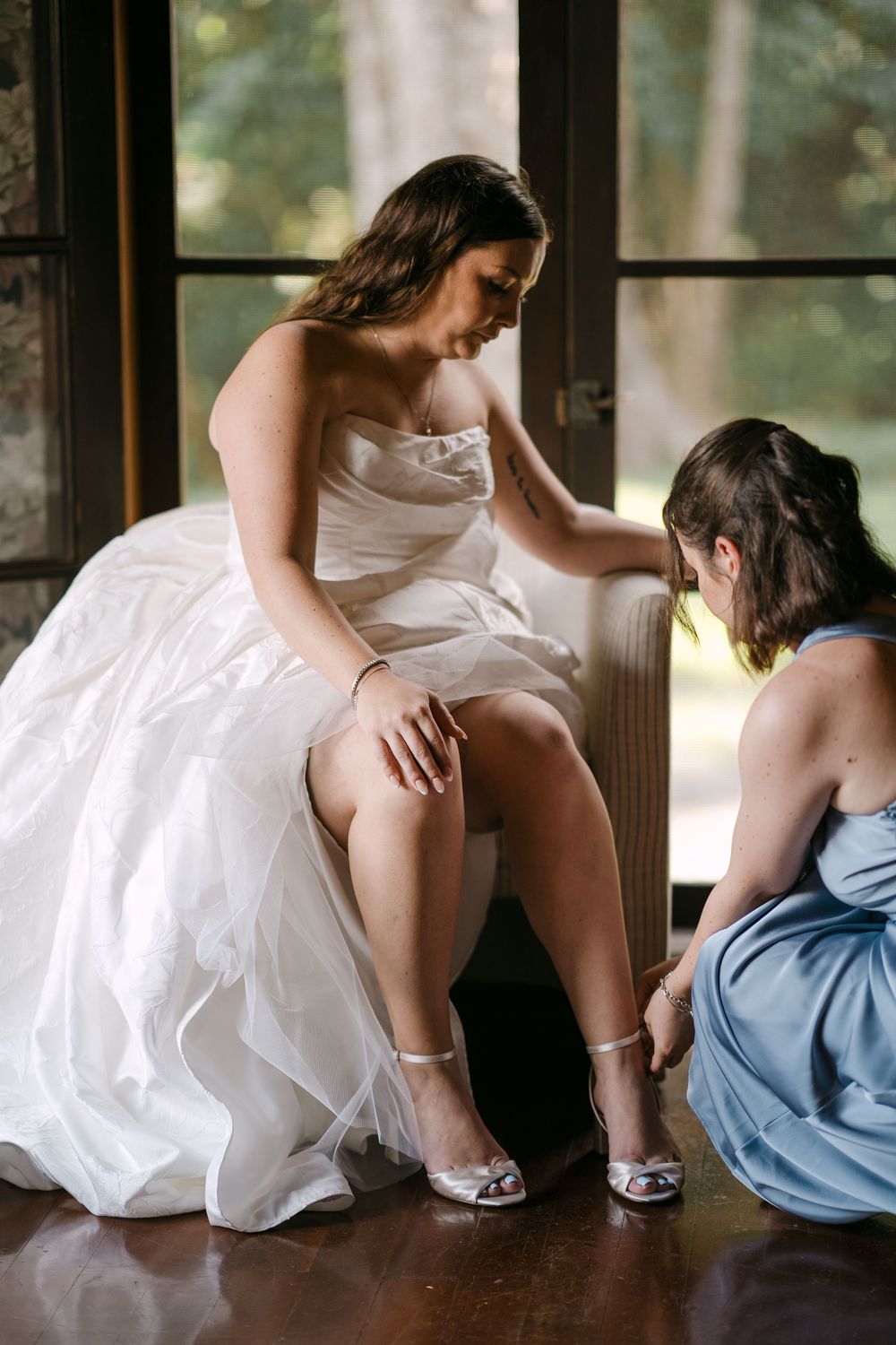 Bridesmaid in blue dress helps bride in white gown put on silver heels before wedding ceremony.