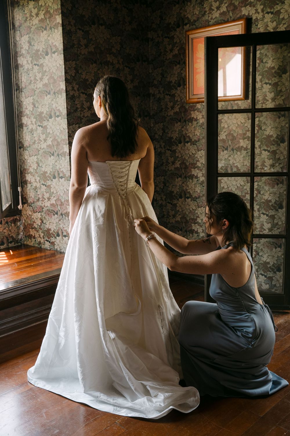 Bridesmaid in blue dress lacing up bride's white wedding gown in elegant room with floral wallpaper.