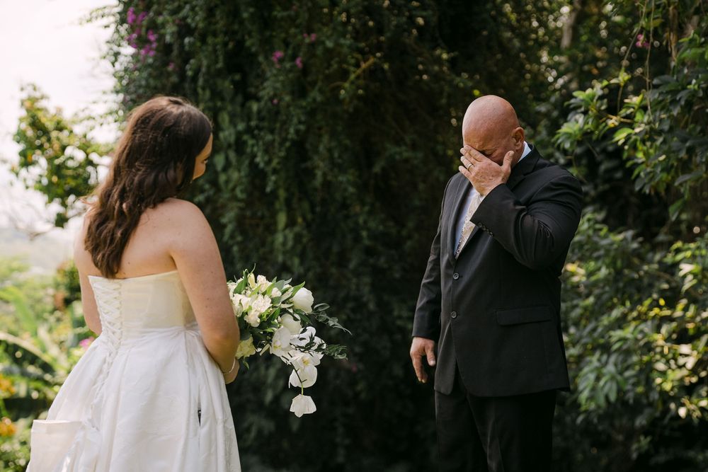 Emotional groom wiping tears during first look with bride holding white bouquet in lush garden wedding.