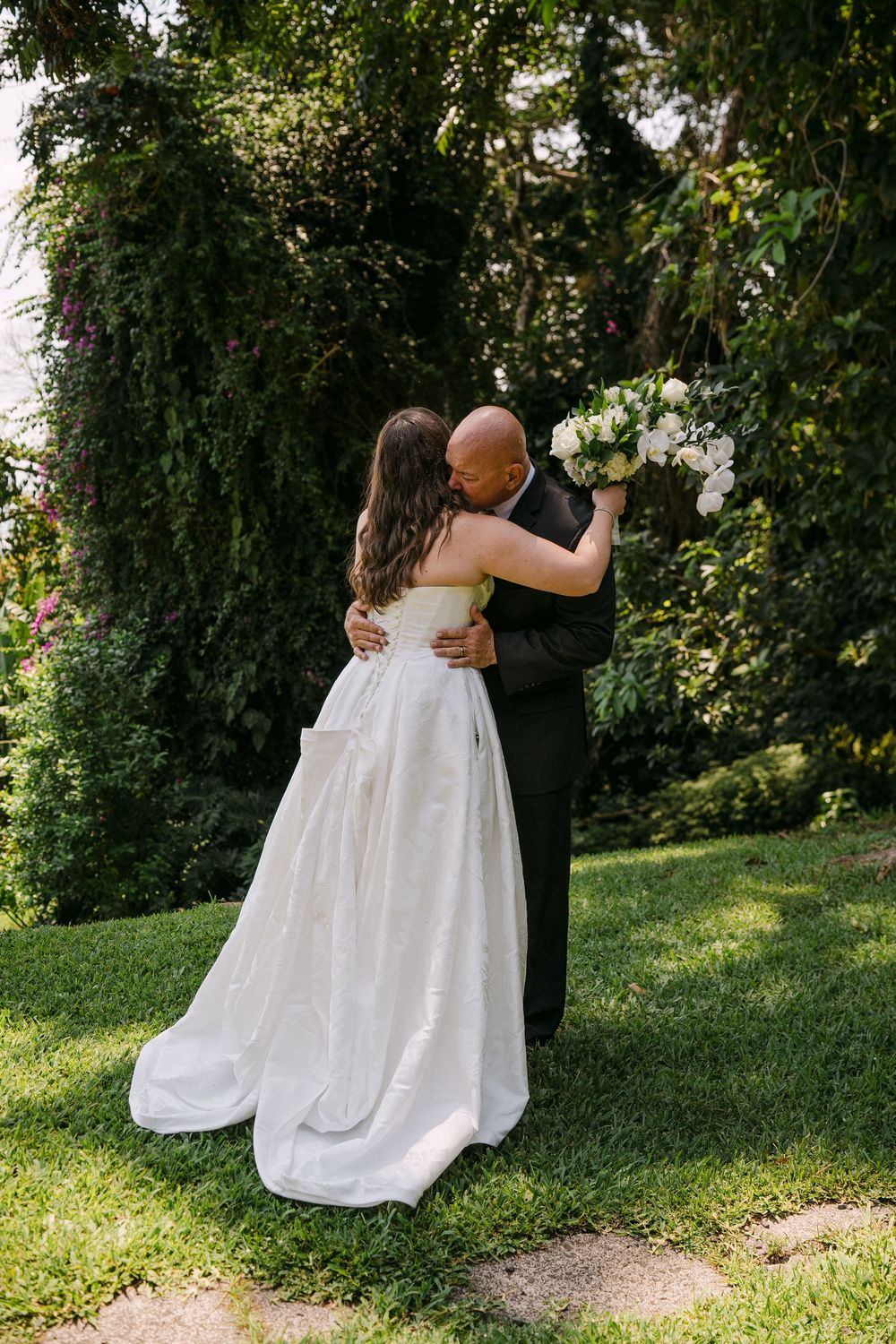 Bride and groom embrace in lush garden, bride in strapless white gown holding white floral bouquet.