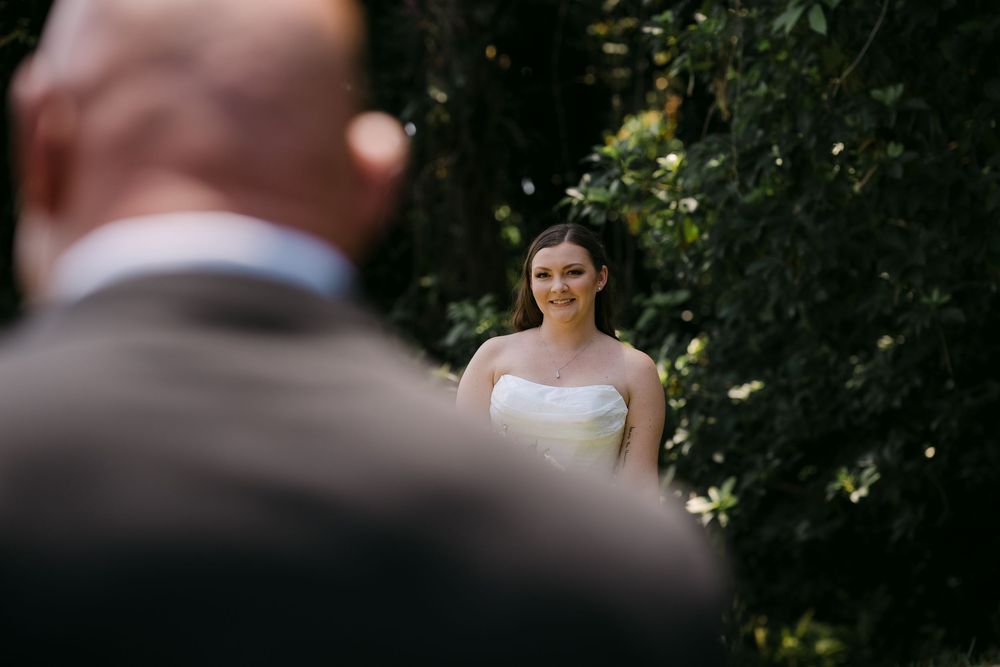 Bride in white strapless gown smiling at groom during outdoor wedding ceremony surrounded by lush greenery.