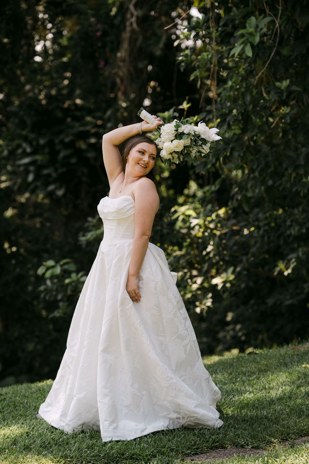 Bride in strapless white ball gown holding white floral bouquet overhead, posing outdoors among lush green trees.