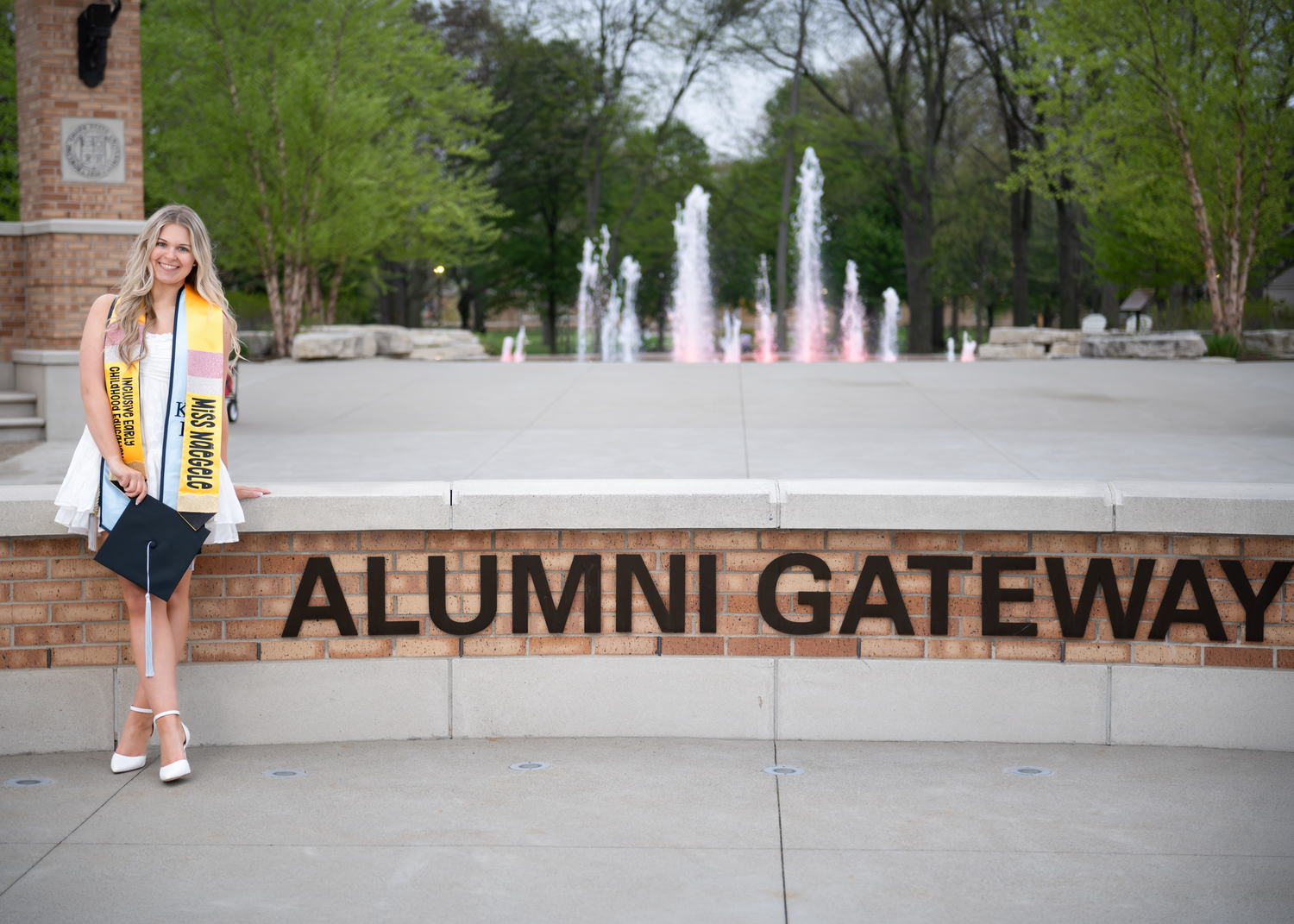 Graduate poses at Alumni Gateway monument with fountains and spring trees in background.