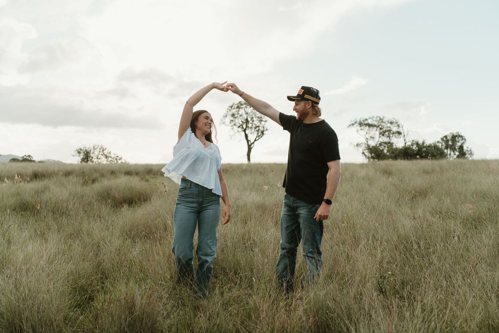 Couple dancing together in a grassy field, holding hands raised, with trees and cloudy sky in the background.