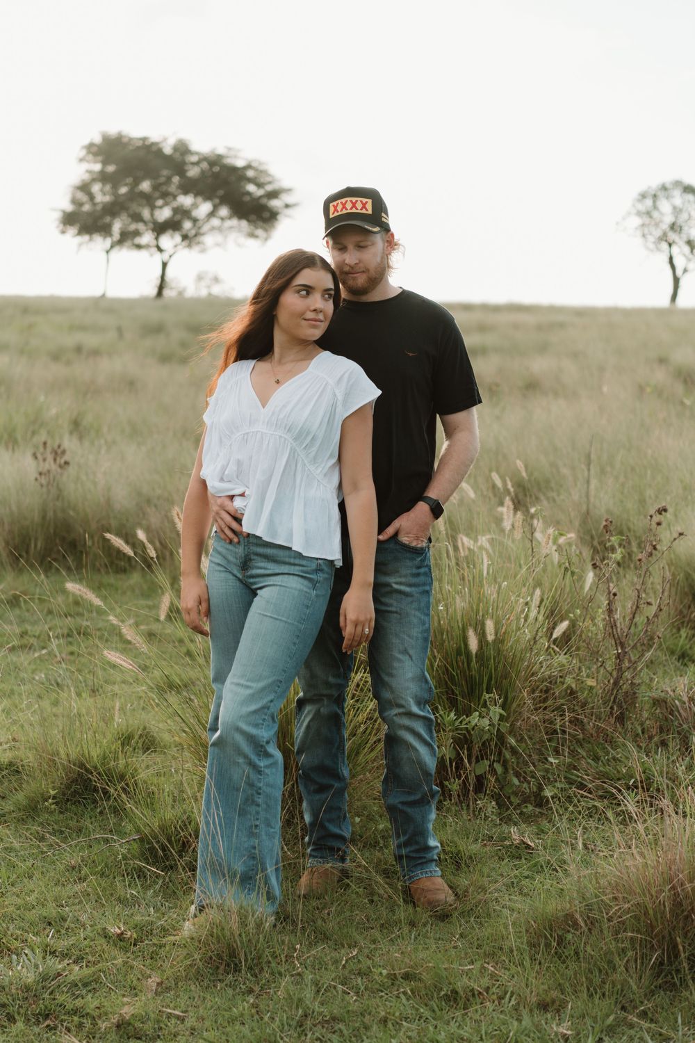 A couple poses together in a grassy field, woman in white top and jeans, man in black shirt and cap.