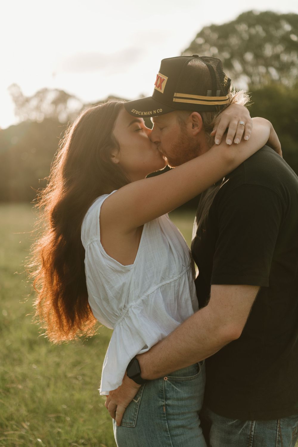 A couple sharing a romantic kiss outdoors at golden hour, woman in white top embracing man in black shirt and cap.