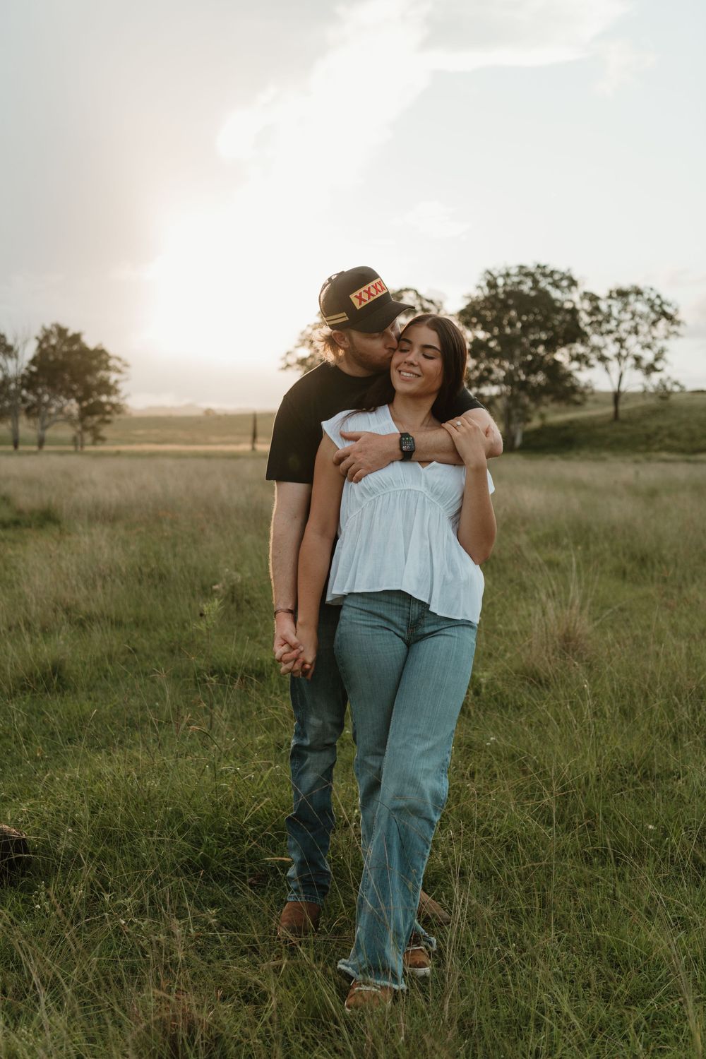 A couple shares a romantic moment in a grassy field at sunset, man kissing womans head while holding hands.