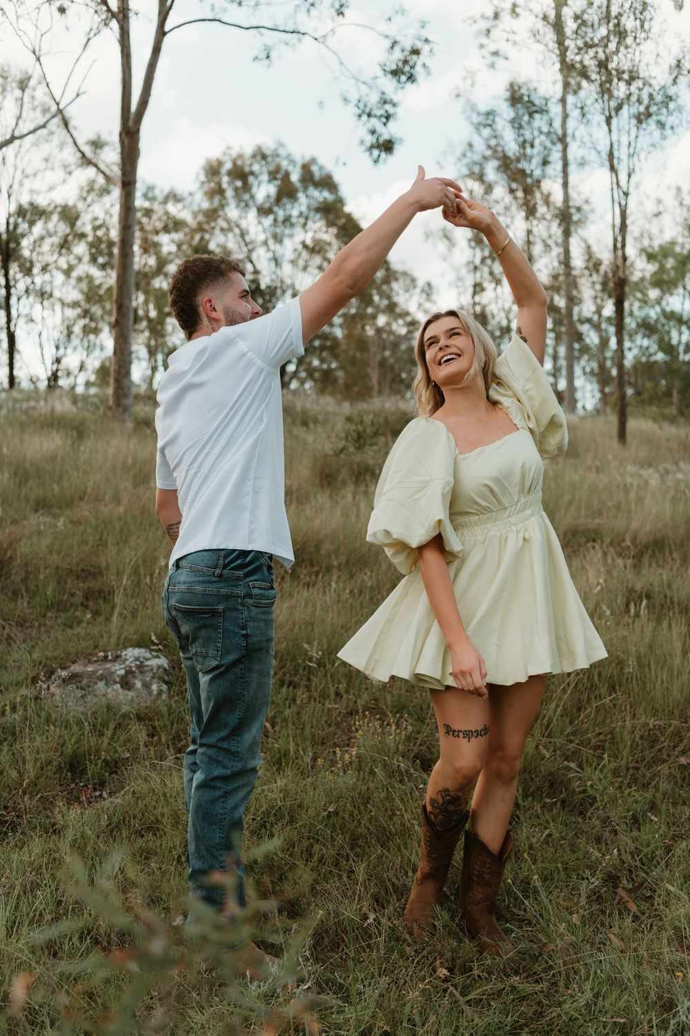 A couple dances playfully together in a grassy field at sunset wearing casual attire and boots. in Boonah Queensland