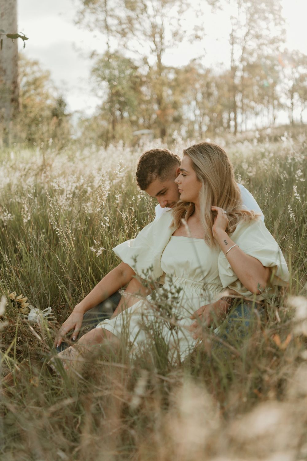 A couple shares intimate moments while sitting in a sun-drenched field of tall grass and wildflowers. in Boonah Queensland