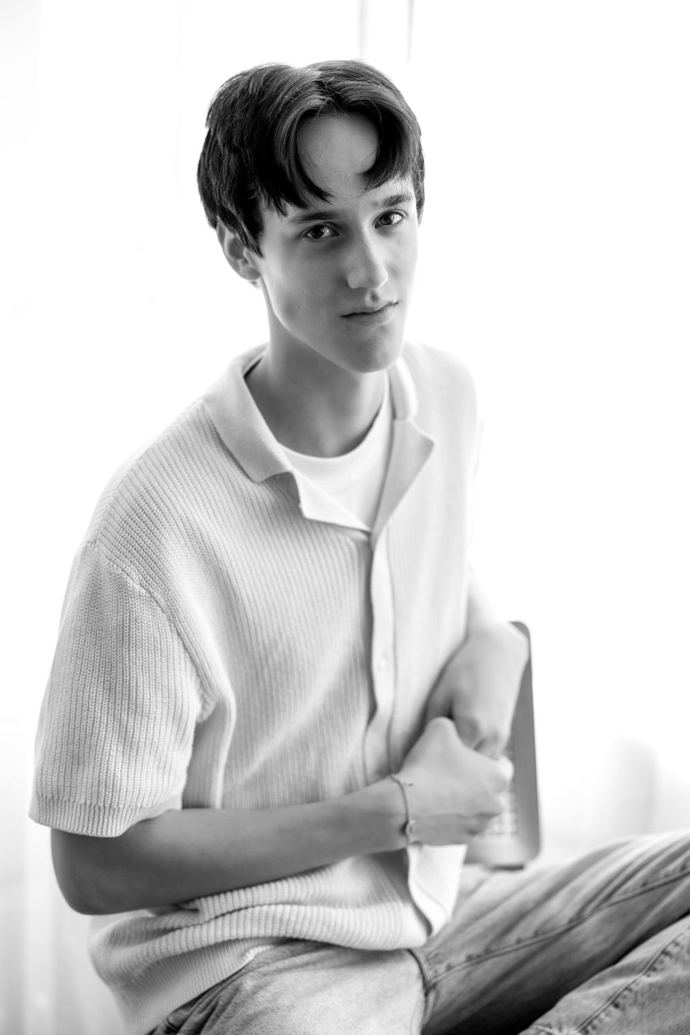 Black and white portrait of a young man in a knit polo shirt sitting near a bright window.
