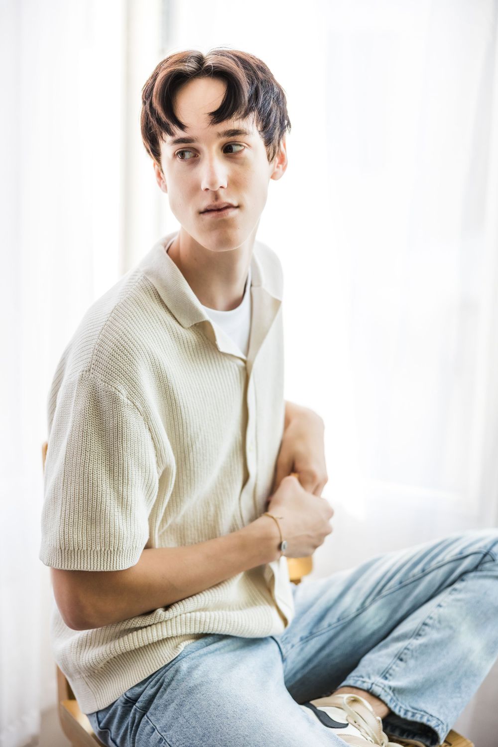 Young man in a light knit polo shirt and jeans sitting casually near a bright white curtained window.