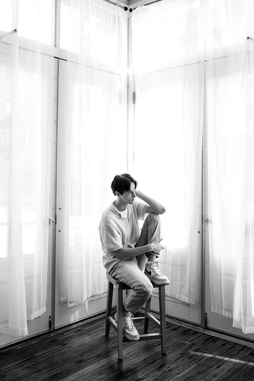 Black and white photo of a young man sitting on a stool in a bright windowed corner room.