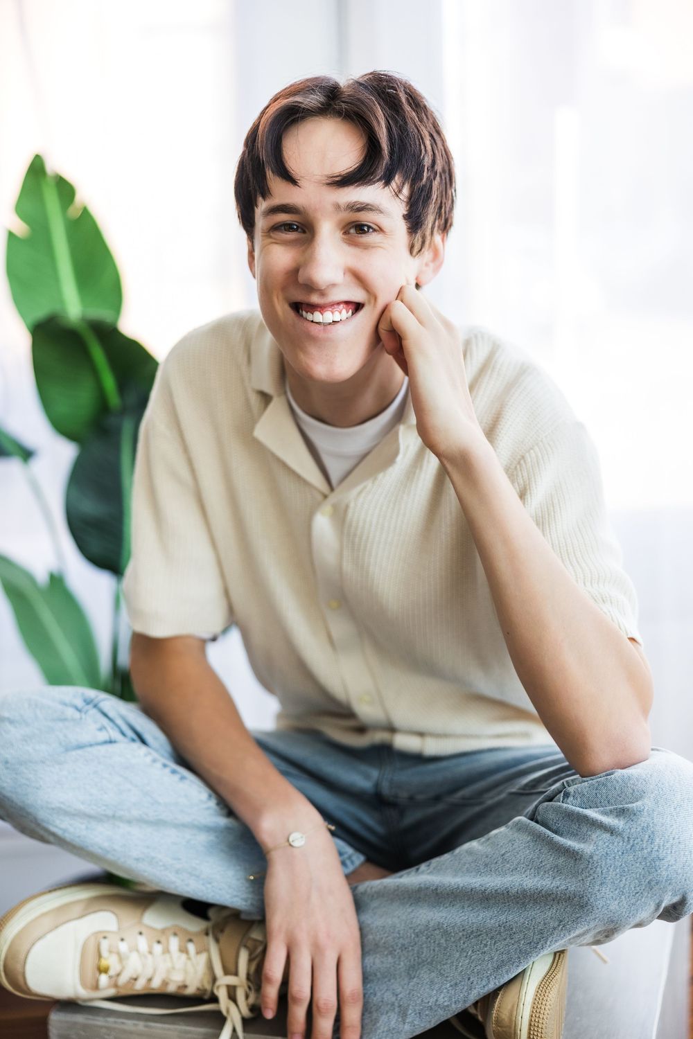 Smiling young man in beige shirt and jeans sitting cross-legged with hand on cheek near indoor plant.