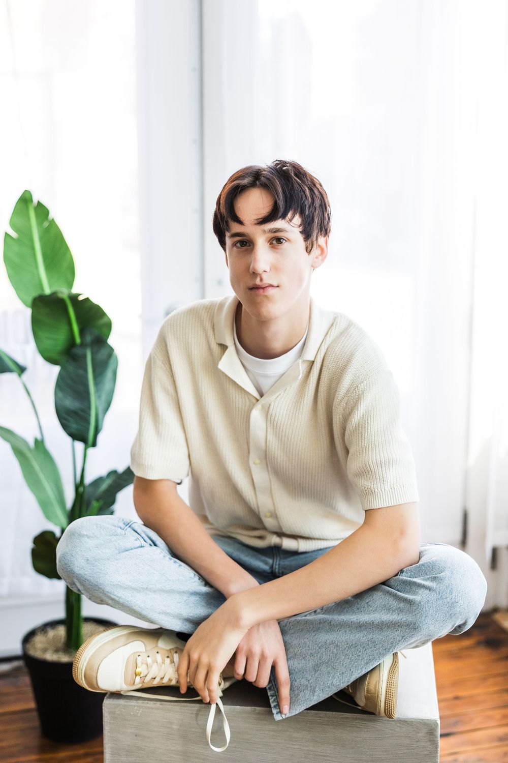Young man in beige ribbed shirt and jeans sitting cross-legged on a box near an indoor plant.