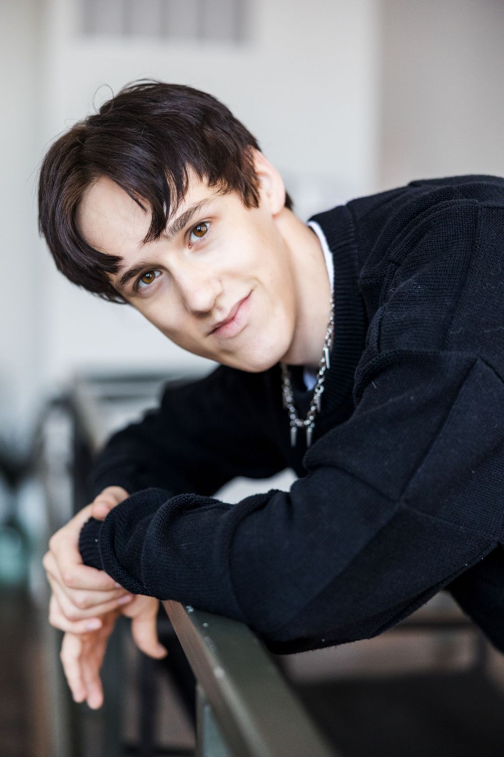 Young man in black sweater with chain necklace leaning on a railing with a relaxed expression.