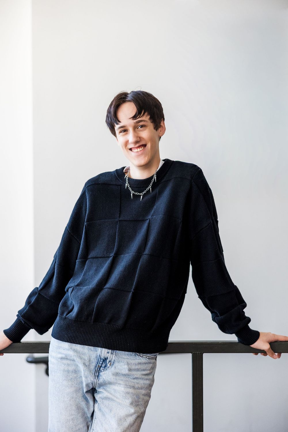 Smiling young man in black sweater and jeans leaning against an indoor railing.