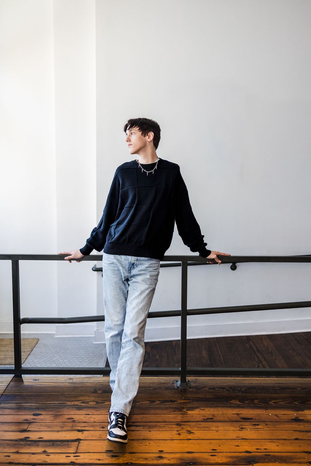 Young man in black sweatshirt and light jeans leaning on indoor railing looking to the side.